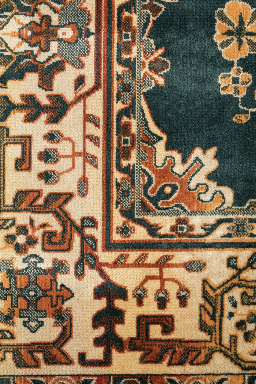 Nahaufnahme eines traditionellen Teppichs mit komplexen Mustern in Rot-, Beige- und Grüntönen. Die Perspektive zeigt eine detaillierte Ansicht der Ecke des Teppichs.