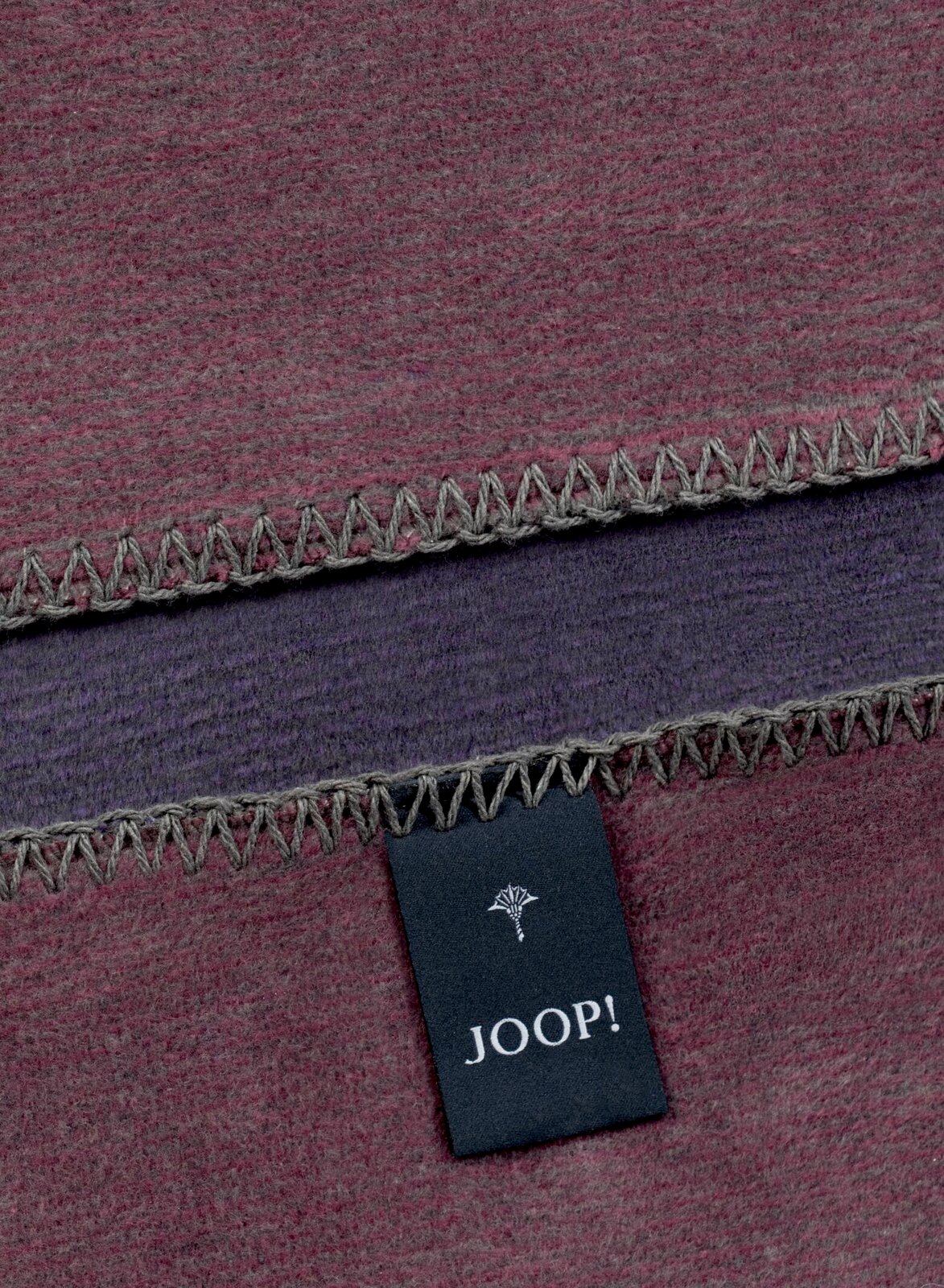 Nahaufnahme einer zweifarbigen Decke mit grauem Zickzackstich und einem Joop!-Etikett.