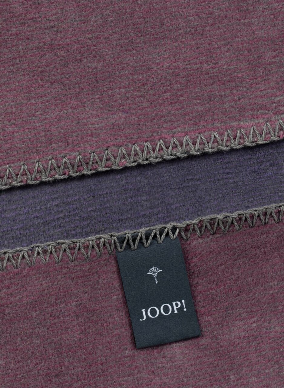 Close-up van een tweekleurige deken met grijze zigzagsteek en een Joop!-label.