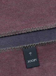 Close-up van een tweekleurige deken met grijze zigzagsteek en een Joop!-label.