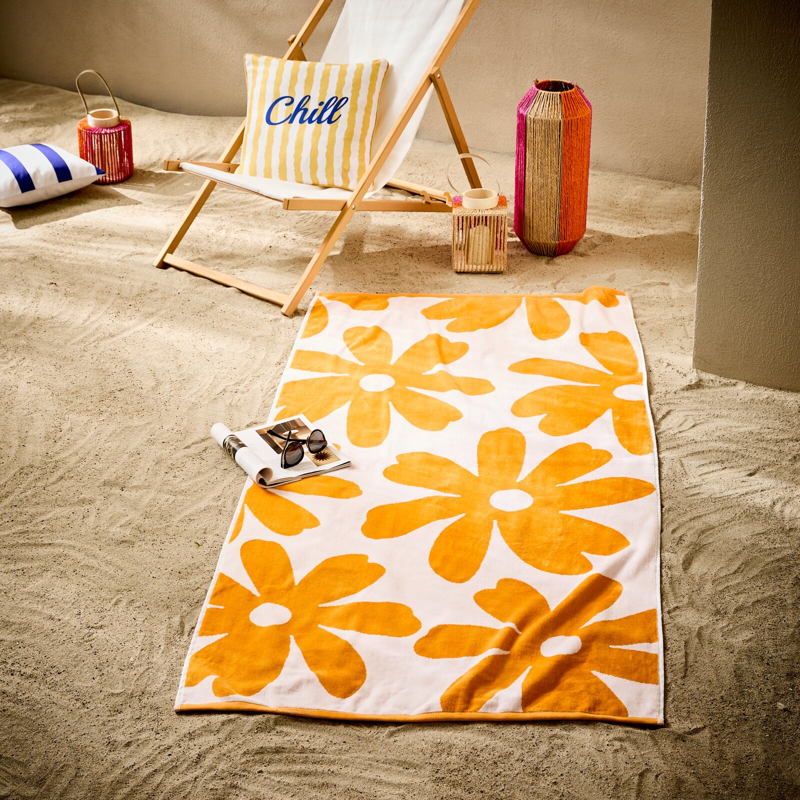 Weißes Strandtuch mit großen orangefarbenen Blumenmotiven auf Sand, im Vordergrund aus leicht schräger Draufsicht fotografiert; daneben Sonnenbrille und Magazin, im Hintergrund Liegestuhl mit Kissen und Deko-Laternen.