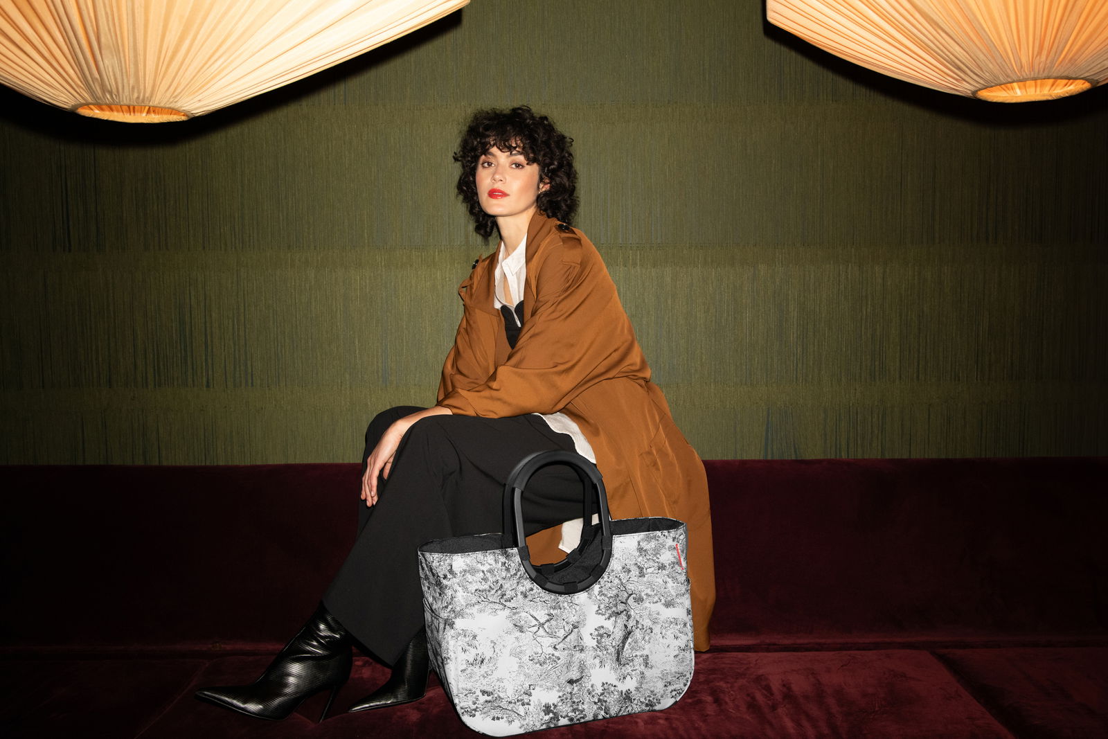 Eine Frau sitzt auf einem Sofa in einem eleganten Raum. Neben ihr steht eine große, stilvolle Tasche mit einem schwarz-weißen Muster und runden Griffen. Die Perspektive zeigt die Tasche von vorne.