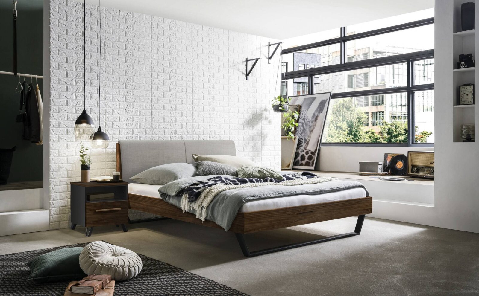 Modernes Schlafzimmer mit einem stilvollen Bettgestell aus Holz und Metall, seitliche Perspektive. Das Bett ist mit grauer Bettwäsche und dekorativen Kissen ausgestattet, umgeben von einer weißen Ziegelwand und großen Fenstern, die viel Licht hereinlassen