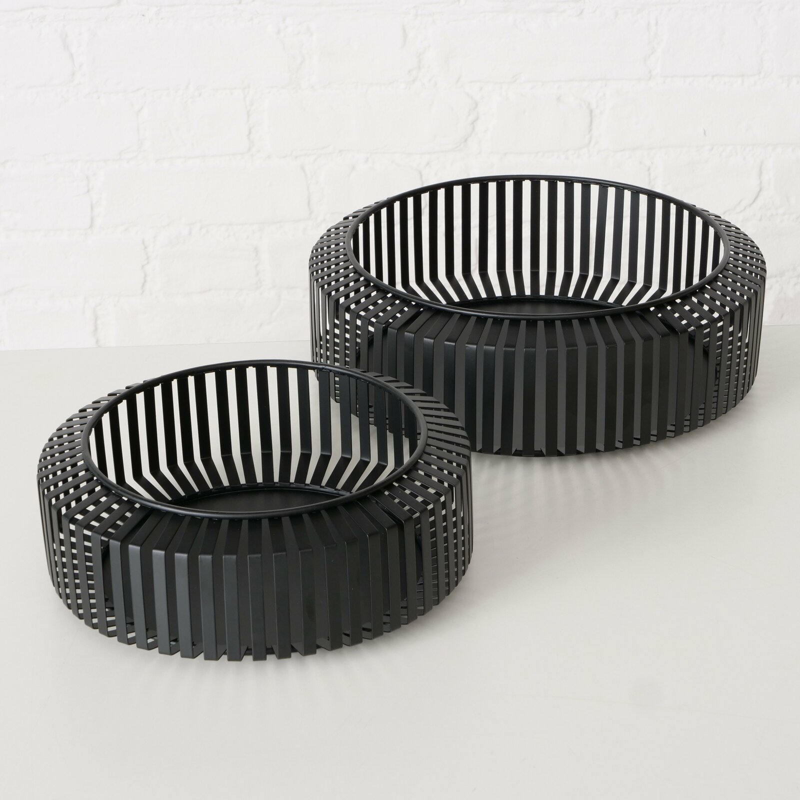 Zwei schwarze, runde Schalen aus Metall mit vertikalen Streben, auf einer weißen Oberfläche vor einer Ziegelwand, aus leicht erhöhter Perspektive.