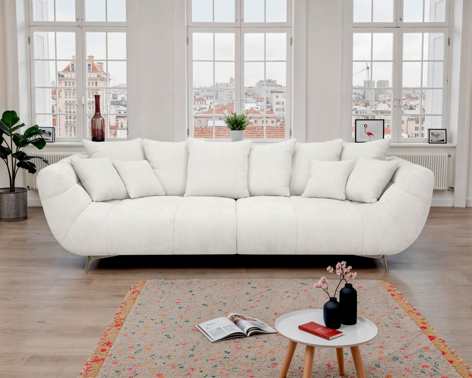 Helles, cremeweißes Stoffsofa mit weicher Polsterung, abgerundeten Armlehnen und mehreren Rückenkissen auf schlanken Metallfüßen, frontal in einem lichtdurchfluteten Wohnzimmer vor großen Sprossenfenstern fotografiert.