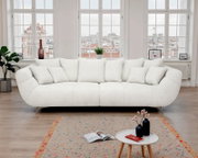 Helles, cremeweißes Stoffsofa mit weicher Polsterung, abgerundeten Armlehnen und mehreren Rückenkissen auf schlanken Metallfüßen, frontal in einem lichtdurchfluteten Wohnzimmer vor großen Sprossenfenstern fotografiert.