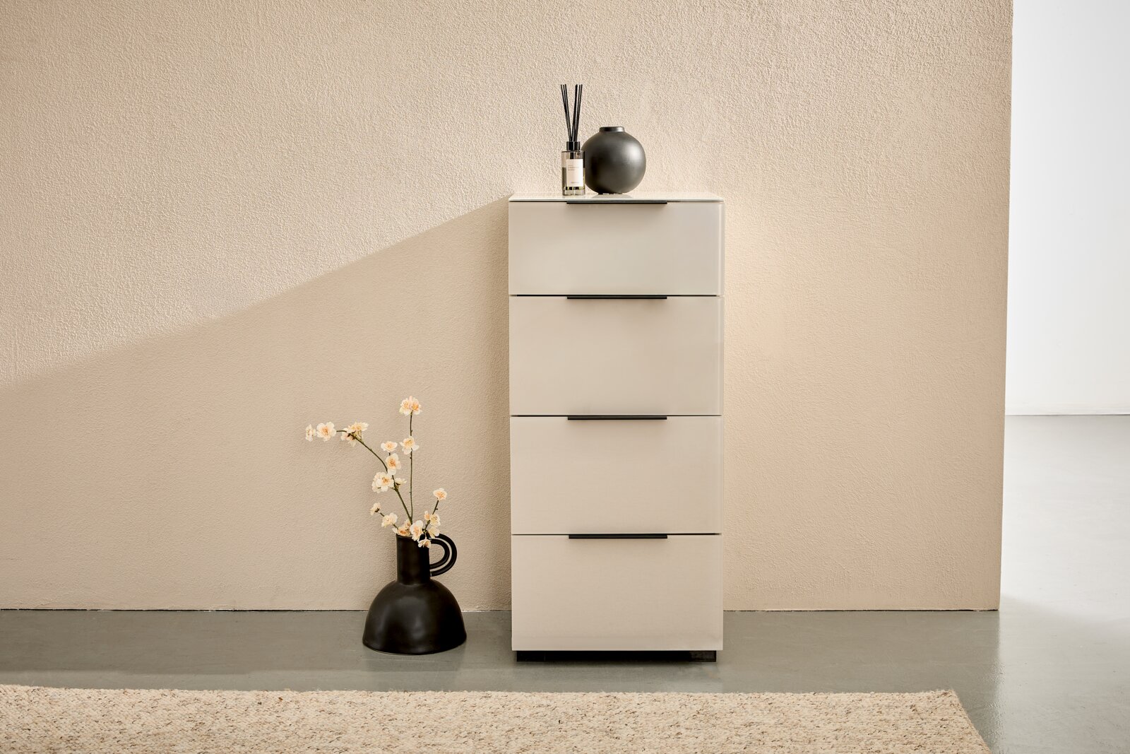 Novel Hoge ladekast MALIN 2.0 Smalle, moderne ladekast in crème/beige met vier lades en zwarte handgrepen, van voren gefotografeerd op ooghoogte voor een gestructureerde muur; bovenop decoratie met een geurverspreider en een zwarte vaas, ernaast een zwarte vloervaas met bloemen.