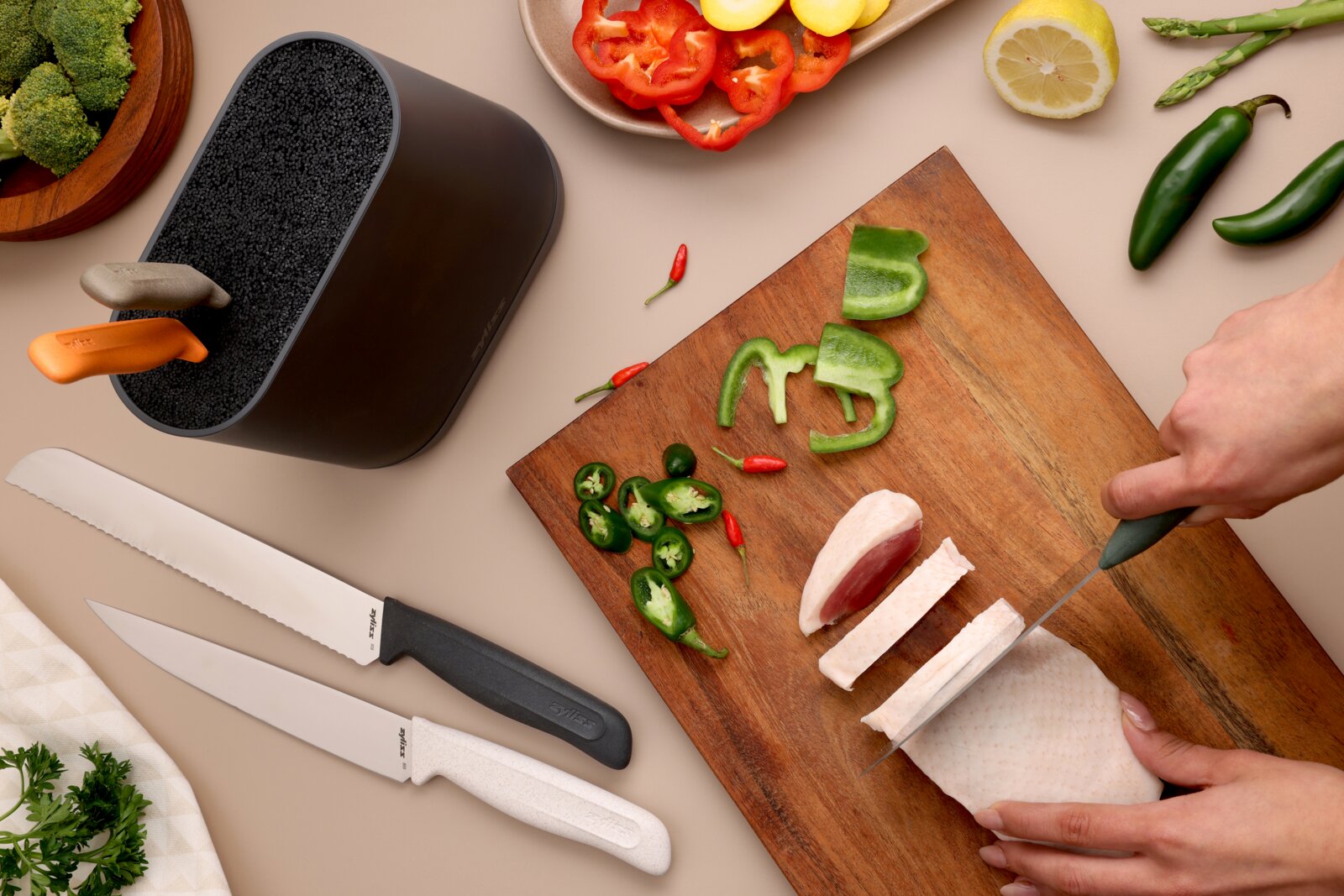 Bovenaanzicht van twee keukenmessen met zwart en wit handvat naast een messenblok, terwijl iemand kip snijdt op een houten snijplank met paprika en jalapeño eromheen.