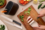 Bovenaanzicht van twee keukenmessen met zwart en wit handvat naast een messenblok, terwijl iemand kip snijdt op een houten snijplank met paprika en jalapeño eromheen.