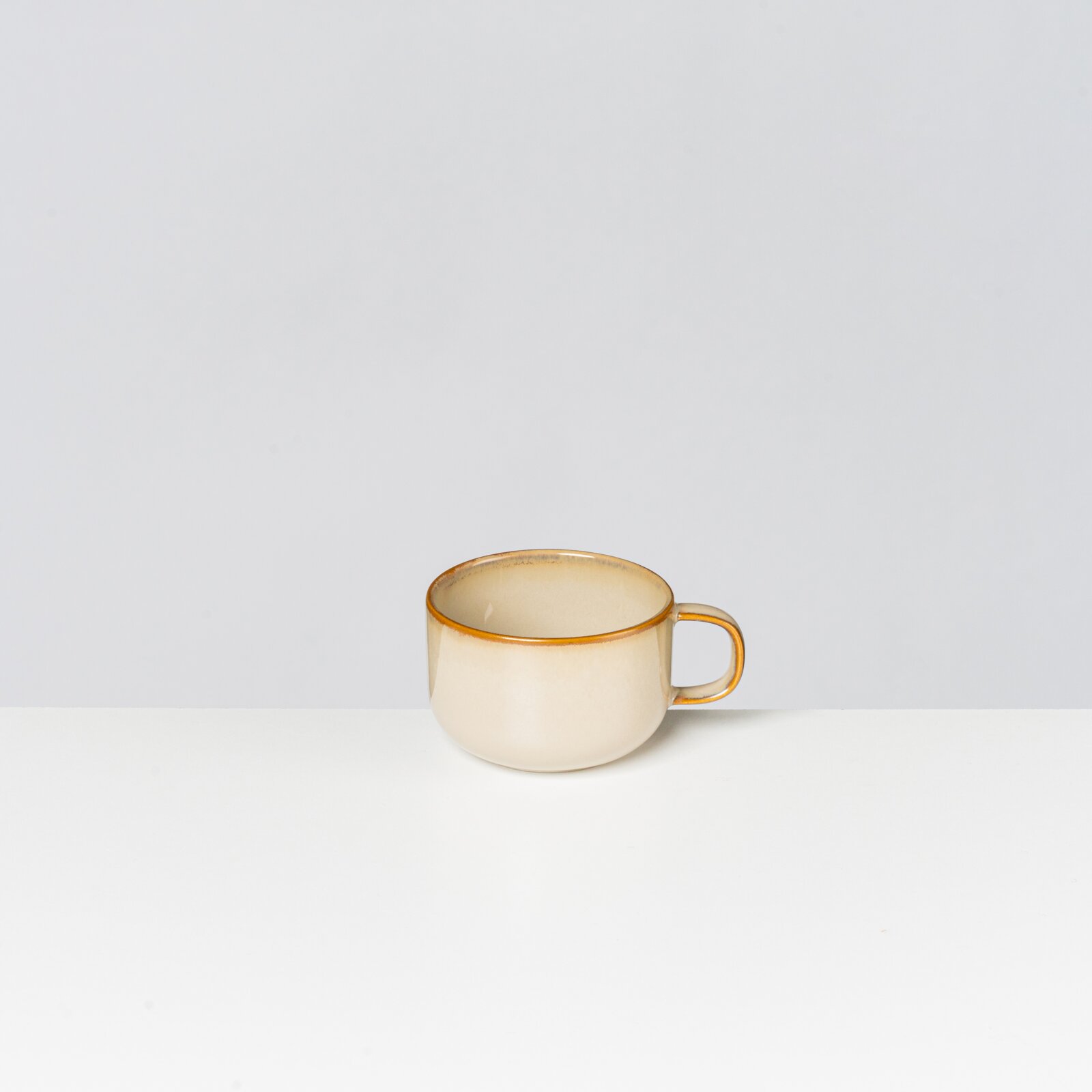Beige Keramik-Tasse mit goldenem Rand und Henkel, freigestellt vor hellem Hintergrund, seitliche Frontansicht leicht von oben