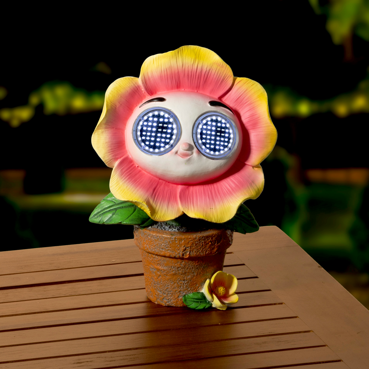 Niedliche Solarfigur in Form einer Blume mit leuchtenden Augen, in einem Blumentopf auf einem Holztisch, frontal fotografiert.