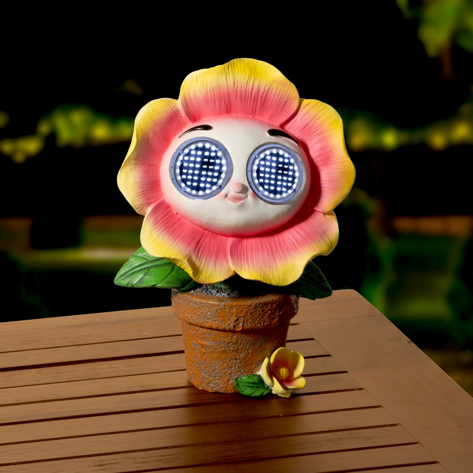 Niedliche Solarfigur in Form einer Blume mit leuchtenden Augen, in einem Blumentopf auf einem Holztisch, frontal fotografiert.