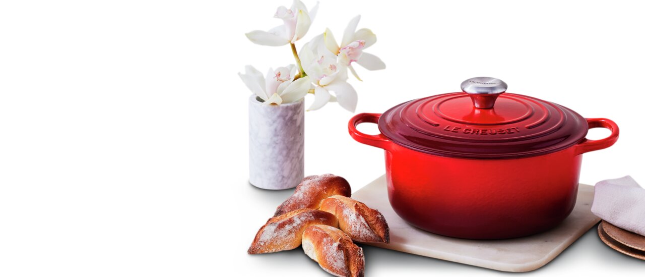 LE CREUSET Bräter SIGNATURE Runder roter Bräter von Le Creuset mit Deckel, seitliche Perspektive, auf einem Tisch mit Blumen und Brot.