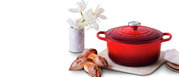 Runder roter Bräter von Le Creuset mit Deckel, seitliche Perspektive, auf einem Tisch mit Blumen und Brot.