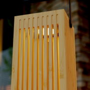Nahaufnahme einer modernen Hängelampe mit rechteckigem Holz-/Bambus-Lampenschirm mit vertikalen Schlitzen und warm leuchtendem Leuchtmittel, fotografiert aus leicht schräger Untersicht (Perspektive von unten)