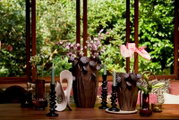 Frontaal aanzicht van een sfeervol gedekte houten tafel met twee sculpturale bruine vazen, roze bloemen, sierlijke zwarte kandelaars en decoratieve woonaccessoires tegen een groene tuinachtergrond.