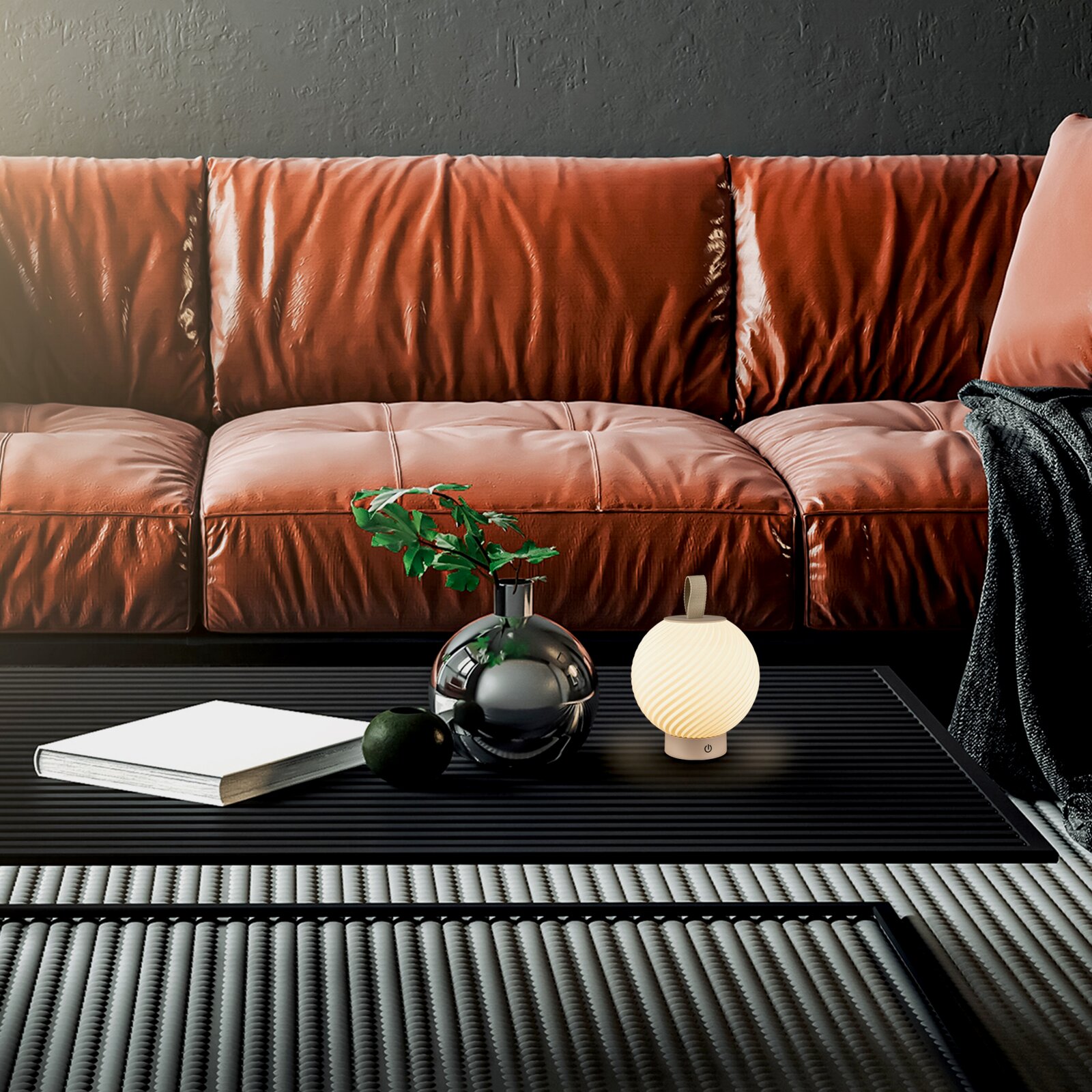 Modernes Wohnzimmer-Setting mit braunem Ledersofa im Hintergrund und schwarzem Couchtisch im Vordergrund; darauf eine runde Glasvase mit grünen Blättern, ein Buch und eine kleine geriffelte Kugel-Tischlampe, frontal aus leicht niedriger Perspektive aufgen