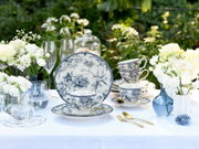 Blau-weißes Porzellan-Geschirr mit floralem Muster (Tassen, Untertassen und Teller) auf festlich gedecktem Tisch im Garten, umgeben von weißen Blumen, Glasglocke und goldfarbenem Besteck, aufgenommen in leicht seitlicher Perspektive auf Tischhöhe.