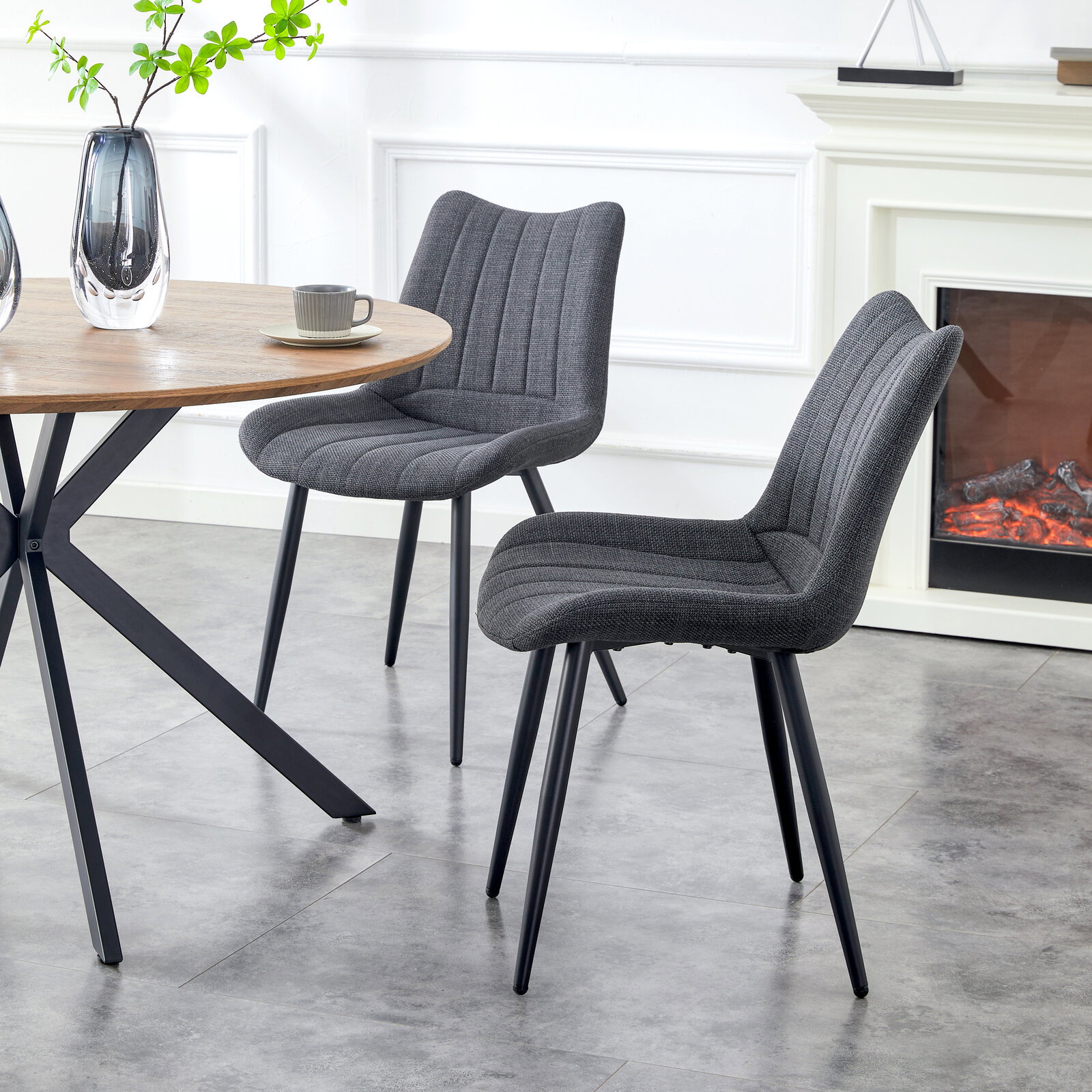 Zwei dunkelgraue, gepolsterte Esszimmerstühle mit Steppnähten und schwarzen Metallbeinen neben einem runden Holztisch in modernem Wohnambiente, seitliche Perspektive auf die Stühle.