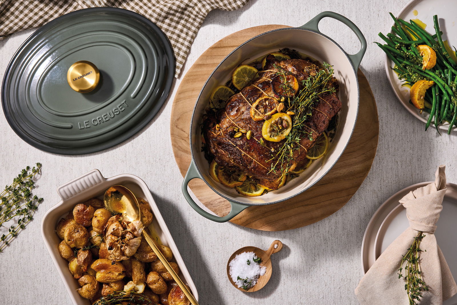 Vogelperspektive auf einen ovalen Bräter von Le Creuset in einem gedeckten Tischarrangement mit gebratenem Fleisch, Kartoffeln und grünen Bohnen.
