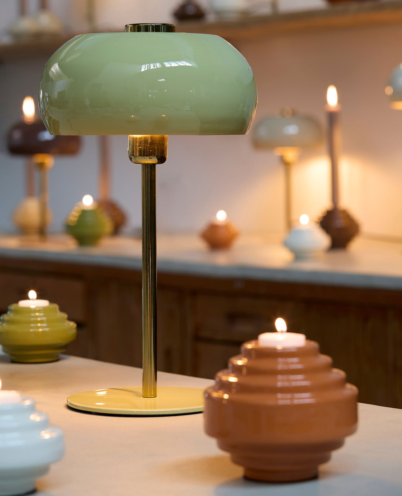 Tischleuchte mit grünem Lampenschirm und goldenem Standfuß, umgeben von Kerzen in einem gemütlichen Raum, seitliche Perspektive.