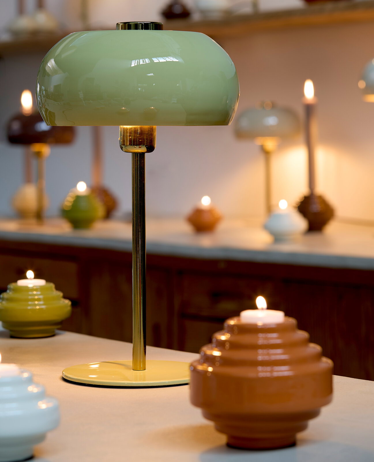 Tischleuchte mit grünem Lampenschirm und goldenem Standfuß, umgeben von Kerzen in einem gemütlichen Raum, seitliche Perspektive.