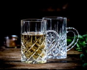 Twee transparante glazen mokken met geslepen ruitmotief, waarvan de voorste gevuld is met een goudkleurige drank, schuin van voren gefotografeerd op een rustieke houten tafel.