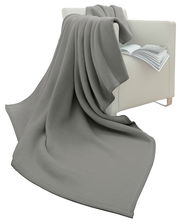 Graue Wohndecke 150 x 200 cm, drapiert über einen weißen Sessel mit einem offenen Buch, seitliche Perspektive