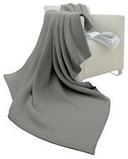 Grijze woondeken 150 x 200 cm, gedrapeerd over een witte fauteuil met een open boek, zijaanzicht