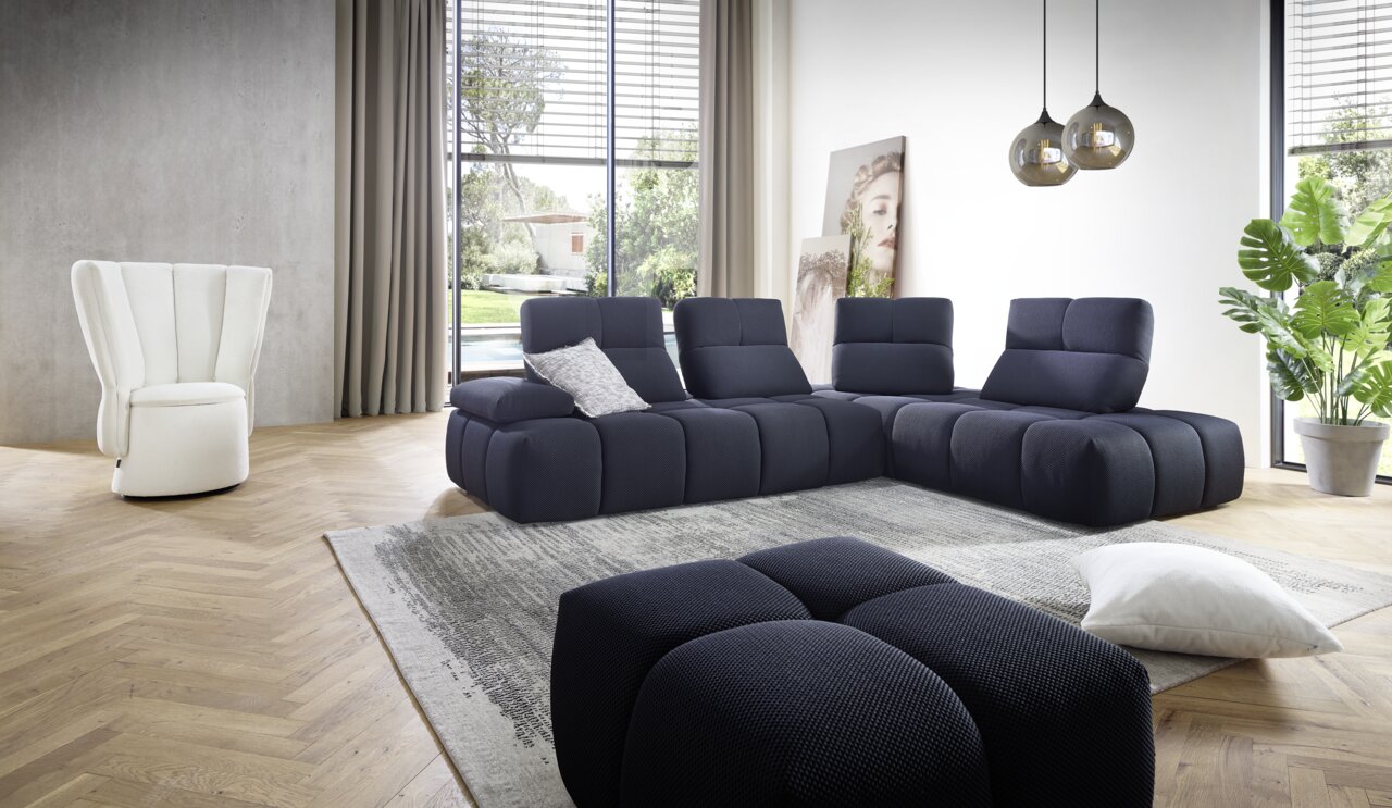 Moderne Eckgarnitur in dunklem Blau, fotografiert aus einer leicht seitlichen Perspektive in einem hellen Wohnzimmer mit großen Fenstern und stilvoller Dekoration.