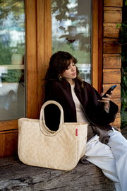 Beige gemusterte Tote-Bag mit ovalen, gepolsterten Henkeln und rotem Markenlabel im Vordergrund, seitliche Frontansicht; im Hintergrund sitzt eine Frau vor einer Holzwand und schaut auf ihr Smartphone.