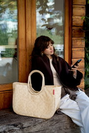 Beige gemusterte Tote-Bag mit ovalen, gepolsterten Henkeln und rotem Markenlabel im Vordergrund, seitliche Frontansicht; im Hintergrund sitzt eine Frau vor einer Holzwand und schaut auf ihr Smartphone.