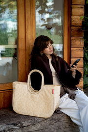 Beige gemusterte Tote-Bag mit ovalen, gepolsterten Henkeln und rotem Markenlabel im Vordergrund, seitliche Frontansicht; im Hintergrund sitzt eine Frau vor einer Holzwand und schaut auf ihr Smartphone.