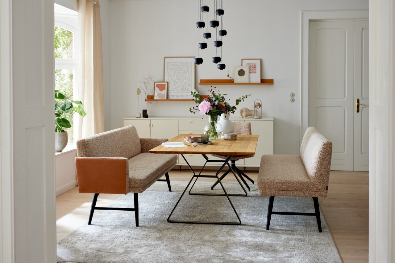 Lichte eethoek met een rechthoekige houten tafel op een zwart metalen onderstel, twee gestoffeerde banken in beige (waarvan één met cognackleurige armleuningen) op een grijs tapijt, met daarboven een zwarte hanglamp; perspectief: frontaal kamerbeeld op oo