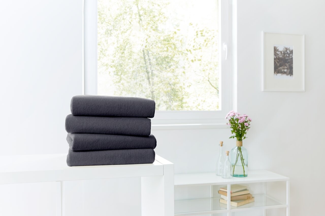 Novel Spannbetttuch TEDDY-FLAUSCH 190x200cm Ein Stapel dunkelgrauer Spannbetttücher auf einem weißen Tisch, im Hintergrund ein Fenster mit Blick ins Grüne. Perspektive von vorne.