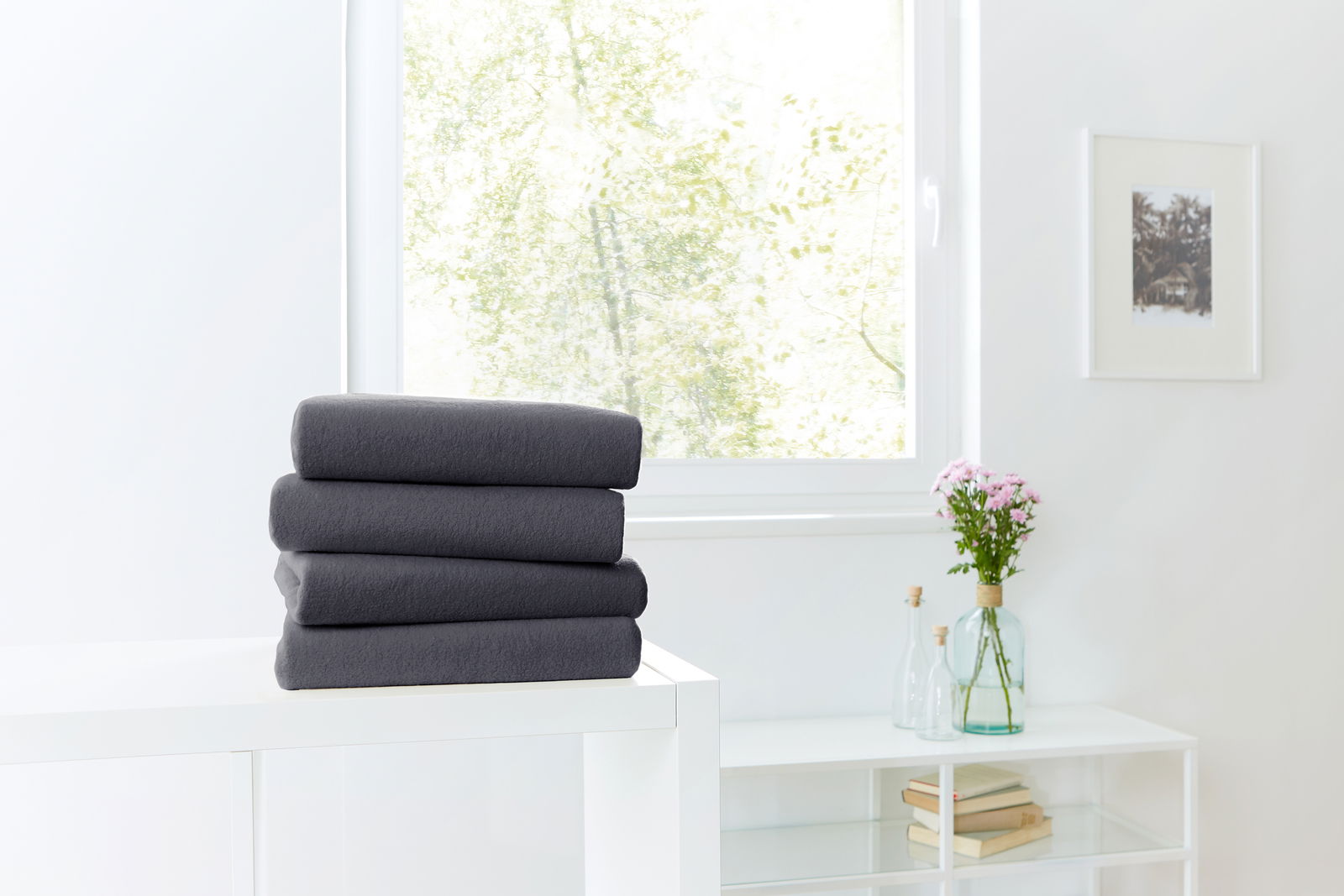 Ein Stapel dunkelgrauer Spannbetttücher auf einem weißen Tisch, im Hintergrund ein Fenster mit Blick ins Grüne. Perspektive von vorne.