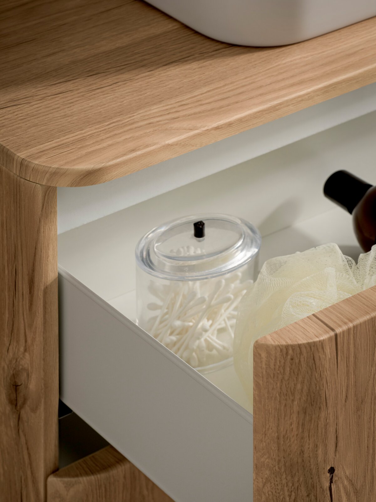 Detailaufnahme aus schräger Seitenperspektive: halb geöffnete weiße Schublade eines Badezimmermöbels mit Holzrahmen, darin transparenter Behälter mit Wattestäbchen und ein Duschschwamm.