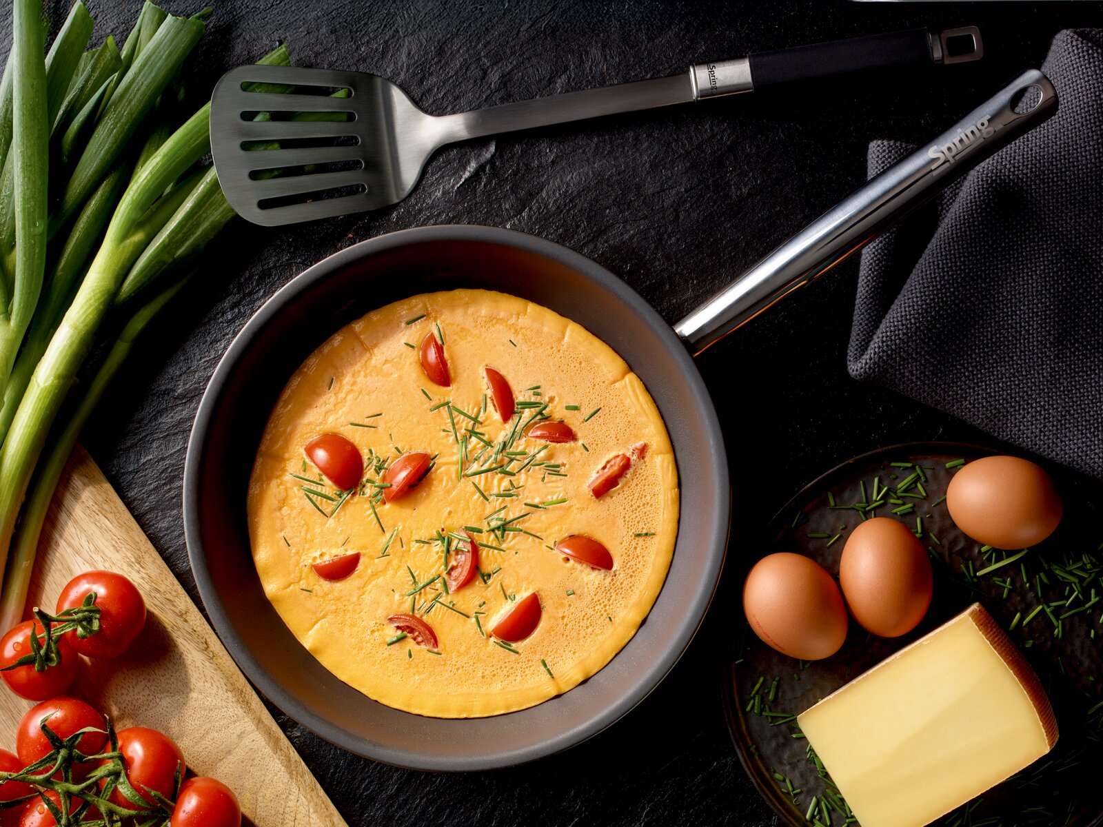 Spring Koekenpan 24 cm BRIGADE ULTIMATE Bovenaanzicht van een koekenpan met omelet, cherrytomaatjes en bieslook op een donkere ondergrond, met eieren, kaas, lente-ui en een spatel ernaast.