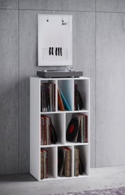 Weißes, schmales Schallplattenregal mit 6 Fächern, gefüllt mit Vinylplatten; oben steht ein Plattenspieler, im Hintergrund ein gerahmtes Poster an einer Betonwand – frontale Ansicht auf das Produkt.