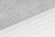 Detailopname vanuit schuin bovenaanzicht van een witte matras met zacht golvend quiltpatroon tegen een lichtgrijze, geweven stoffen achtergrond.