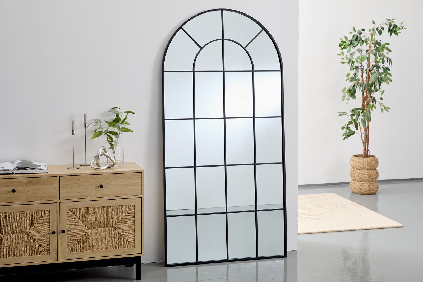 Großer, rechteckiger Spiegel mit schwarzem Rahmen und Gittermuster, an eine weiße Wand gelehnt, neben einer Holzkommode und einer Pflanze. Perspektive von vorne.