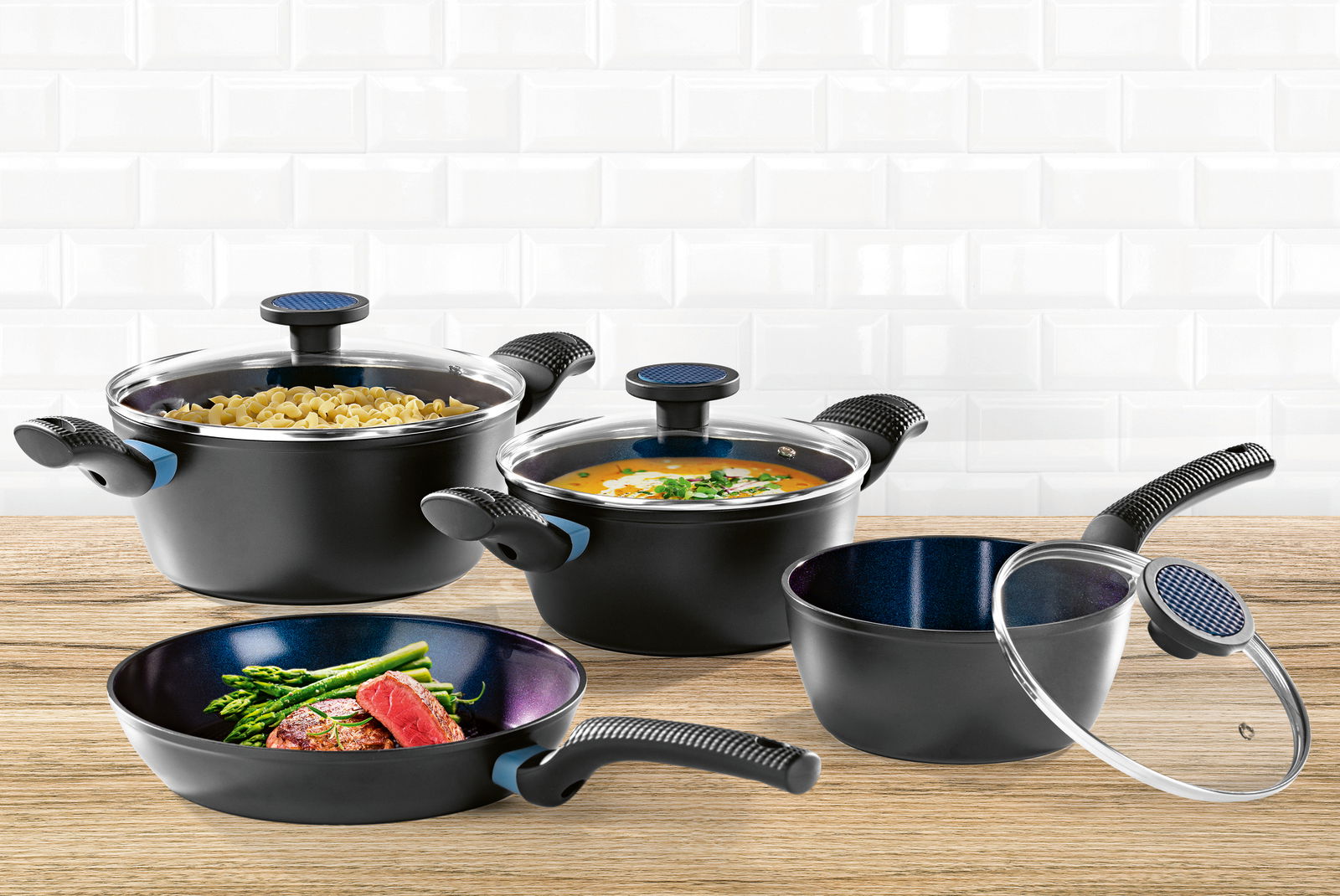 Topfset 7-tlg. in schwarzer Farbe, bestehend aus Töpfen und Pfannen mit Glasdeckeln, auf einer Holzoberfläche vor einer weißen Kachelwand, aus der Vorderperspektive.