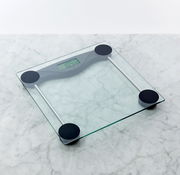 Digitale Personenwaage aus transparentem Glas mit vier schwarzen Standfüßen und LCD-Display, schräg von oben auf einer hellen Marmoroberfläche fotografiert.