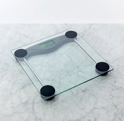 Digitale Personenwaage aus transparentem Glas mit vier schwarzen Standfüßen und LCD-Display, schräg von oben auf einer hellen Marmoroberfläche fotografiert.