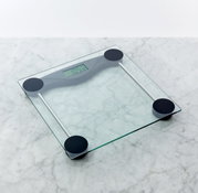 Digitale Personenwaage aus transparentem Glas mit vier schwarzen Standfüßen und LCD-Display, schräg von oben auf einer hellen Marmoroberfläche fotografiert.