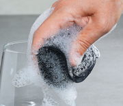 Hand hält einen schwarzen Silikonschwamm, der mit Seifenschaum bedeckt ist, beim Reinigen eines Glases. Perspektive von oben.