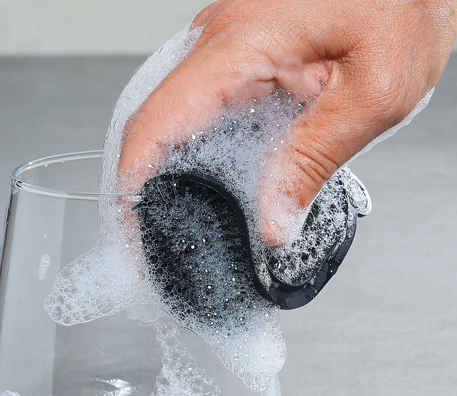 Hand hält einen schwarzen Silikonschwamm, der mit Seifenschaum bedeckt ist, beim Reinigen eines Glases. Perspektive von oben.