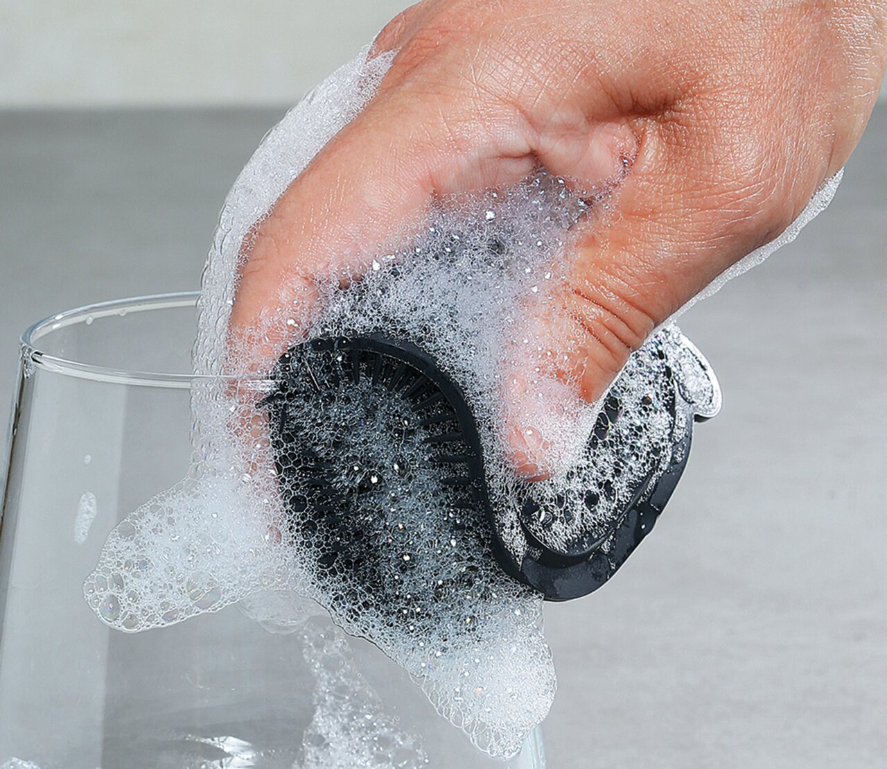 Hand hält einen schwarzen Silikonschwamm, der mit Seifenschaum bedeckt ist, beim Reinigen eines Glases. Perspektive von oben.