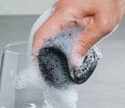 Hand hält einen schwarzen Silikonschwamm, der mit Seifenschaum bedeckt ist, beim Reinigen eines Glases. Perspektive von oben.