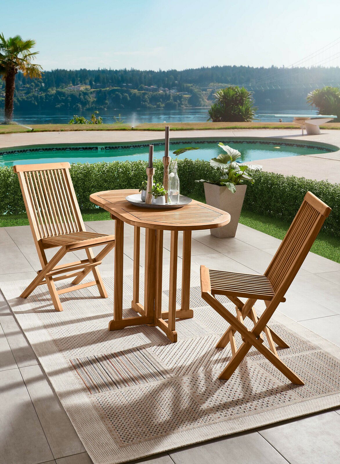 Zwei Holzklappstühle und ein passender Tisch auf einer Terrasse mit Blick auf einen Pool und eine malerische Landschaft im Hintergrund, aus seitlicher Perspektive.